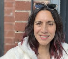 Eelyn Lee, colour portrait, shades on dark hair, sunny day, brick wall background.